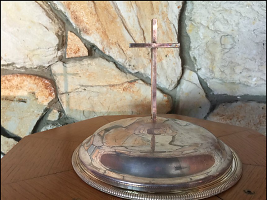 A silver communion vessel with a cross, resting on a wooden table against the stone wall of the sanctuary